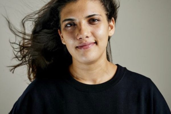 Jessica smiles against a light grey background. They have long dark hair and wear a black t shirt with the National Citizenship Logo on the chest.