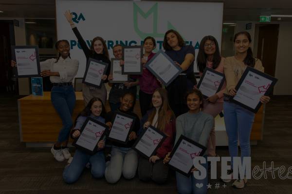 A group of Stemettes Agile Academy attendees holding up their certificates and smiling in front of a QA banner. The image has a dark overlay.