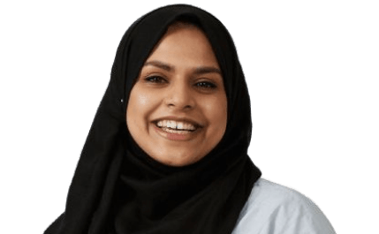 Shajida Akthar wearing a black headscarf smiling in front of a white background.