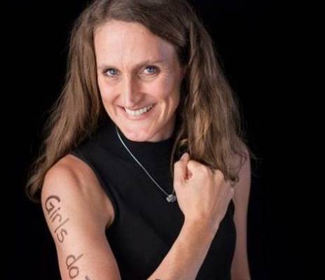 Emma McGuigan with a black vest top smiling in front of a black background with text written on their arm.