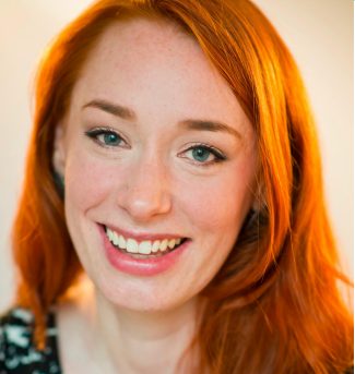 Dr Hannah Fry with long red hair smiling in front of a light coloured background.