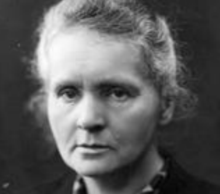Marie Curie wearing a spotted shirt posing in front of a black background.