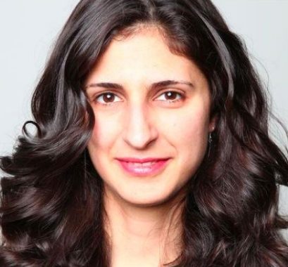 Nina Tandon wearing pink lipstick smiling in front of a grey background.