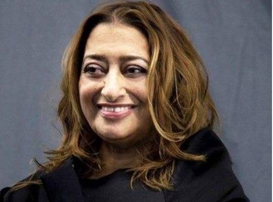 Zaha Hadid wearing a black coat smiling in front of a grey background.