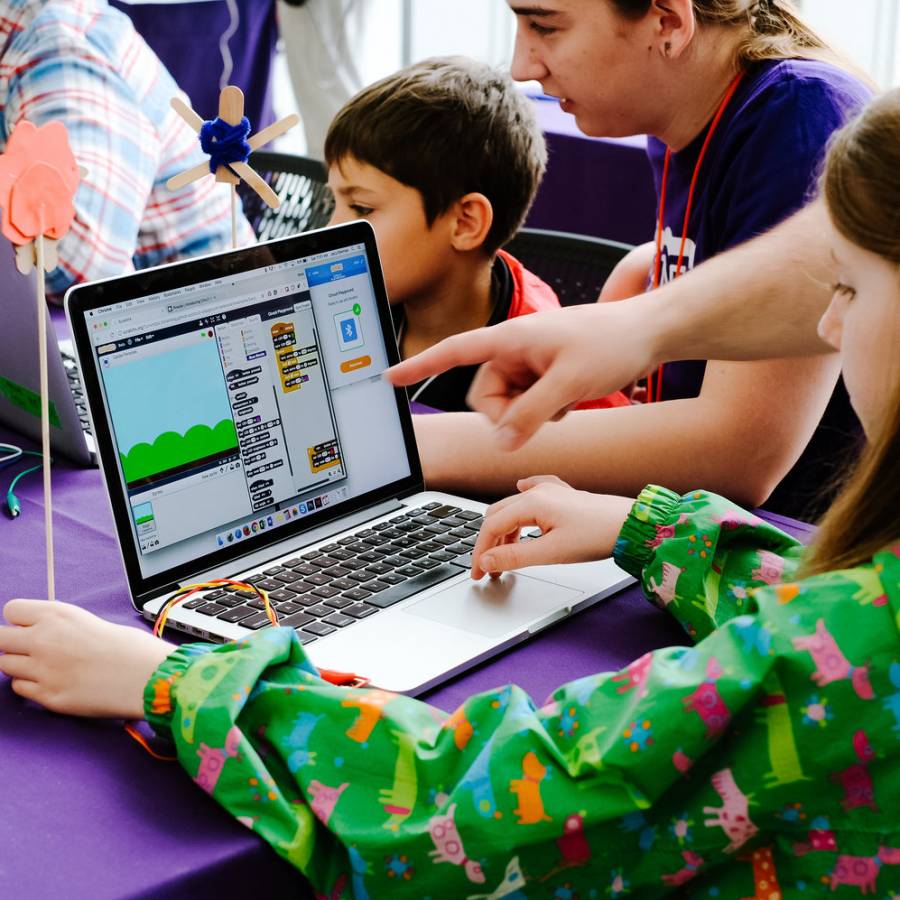 A person working on a laptop trying out a Scratch project. A person is pointing to the screen, helping the individual on the laptop.