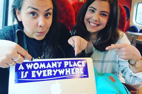 Two people on a train pointing at a laptop sticker that reads 'A womans place is everywhere'.