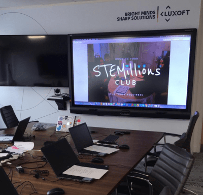 Room with computers and chairs as well as a smart board showing the Stemillions club logo.