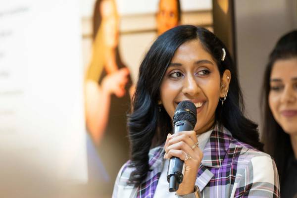An individual in a tartan blazer and a pearl barette in their long black hair. They are holding a microphone and smiling. They are against a background with a blurry presentation and another individual with long brown hair.