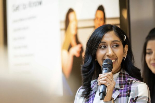 An individual in a tartan blazer and a pearl barette in their long black hair. They are holding a microphone and smiling. They are against a background with a blurry presentation and another individual with long brown hair.