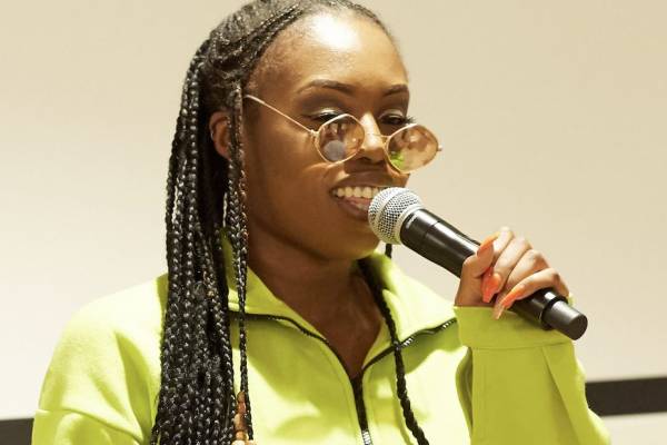 A young person with braids speaking into a microphone against a white background. They are wearing sunglasses with round lenses, and a neon green jumper.