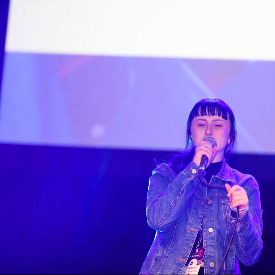 A person wearing a black shirt and a denim jacket stands in front of a large projector on a stage holding a microphone.