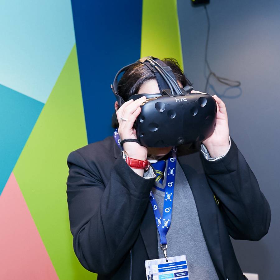 A person standing against a geometric multicolour background. They are wearing a black blazer with a grey shirt underneath and are using a VR headset which they are holding on to with both hands.