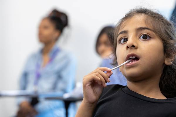 A child is seen listening to something off screen. They have a black t-shirt on, dark hair in a ponytail and a lollypop stick in their mouth. Other people can be seen in the background.
