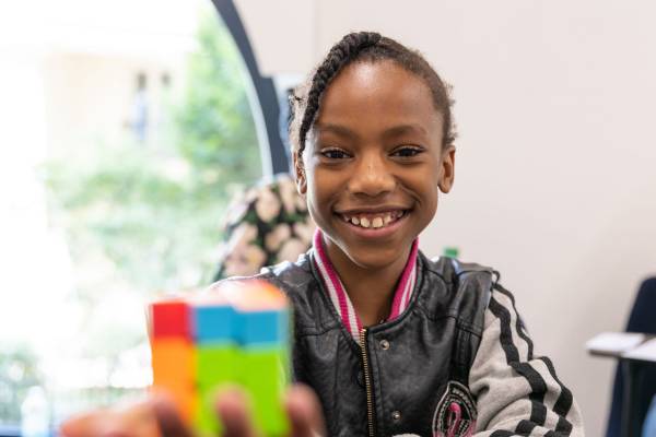 A child with braids is seen smiling and holding out a Rubix cube. There are other children seen in the background.