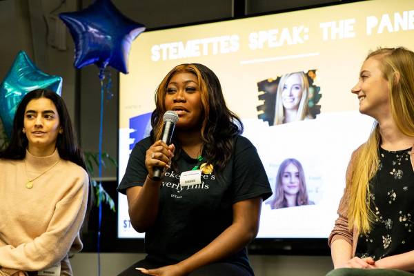 Three Stemettes Alumnae sitting on chairs in a panel formation. The middle person holds a microphone. From left to right they are wearing a beige jumper with long black hair and a gold necklace, a black graphic t shirt with light brown hair and a floral t shirt, pink bomber jacket and blonde hair.
