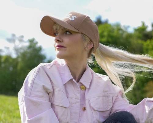 Kim is sitting on grass with trees in the background. She is wearing a brown cap with her long blode ponytail pulled through, a pink denim jacket and black jeans. She is looking to the side and smiling.