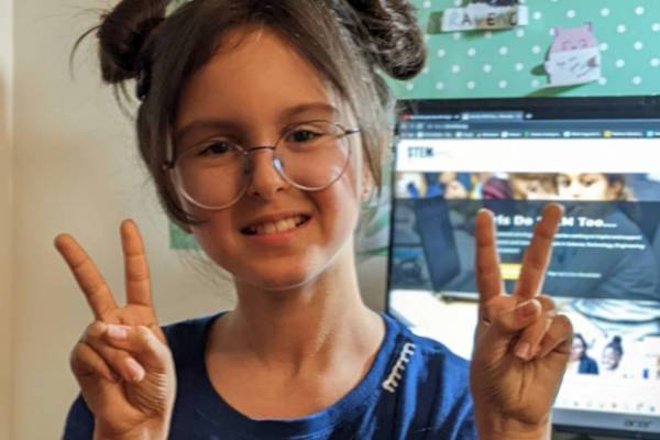 Soraya is posing with two peace signs in front of her computer screen. She has two buns in her hair and wears glasses and a blue t shirt.