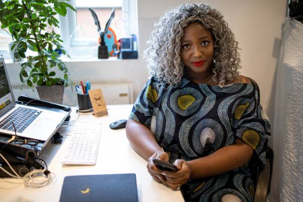 Anne-Marie Imafidon sitting at a desk holding her phone. She is turned to the side and is wearing a printed dress. She has grey hair, red lipstick and is smiling. A laptop can be seen to the left of the image, and a notebook in the foreground.