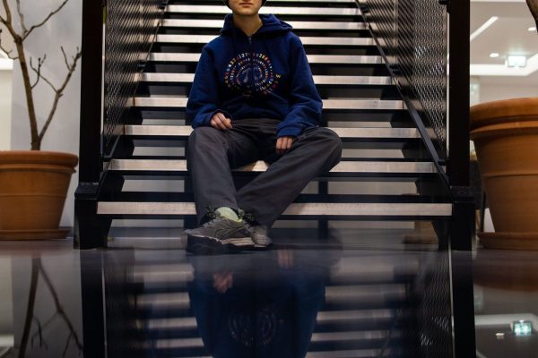 Isabella sits at the bottom of a staircase wearing a Stemettes beanie, a blue hoodie, grey trousers and grey shoes. They are smiling at the camera.