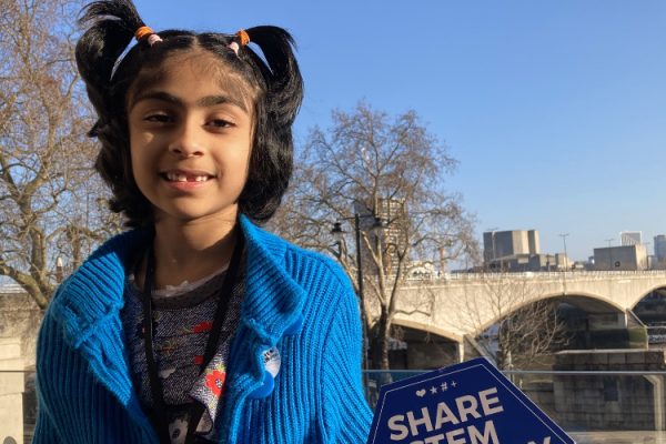 Anoushka smiles at the camera in front of the Waterloo bridge. Her hair is in pigtails and she wears a blue cardigan. She holds a sign says 'Share STEM Herstory'.