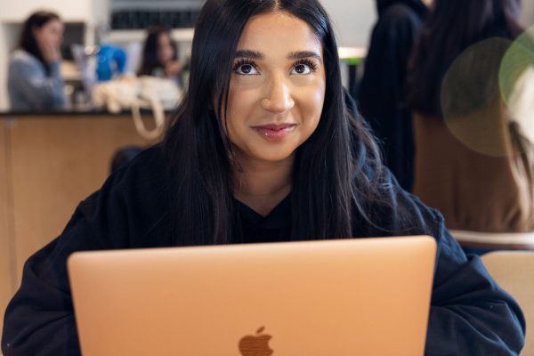 A member of TeamStemette sits and smiles at the camera. Her macbook is infront of her, and other people can be seen behind.