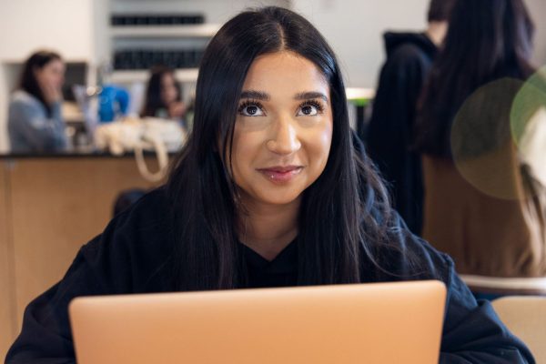 A member of TeamStemette sits and smiles at the camera. Her macbook is infront of her, and other people can be seen behind.