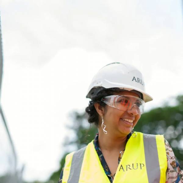 A young women is smiling at someone of the right side of the image. She is on a construction site as she is wearing a high-visibility vest, a hardhat and a pair of safety goggles. She has long, black hair in a bun and is wearing a colourful patterned blouse.