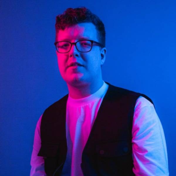 Matt Hardwick looking at the camera. He is wearing a white long sleeved jumper and a black waistcoat. He has short brown hair and is wearing black rounded-square glasses. He is sat in front of a blue background and there is red light shining on him from the left hand side of the picture. The source of the light is not visible.
