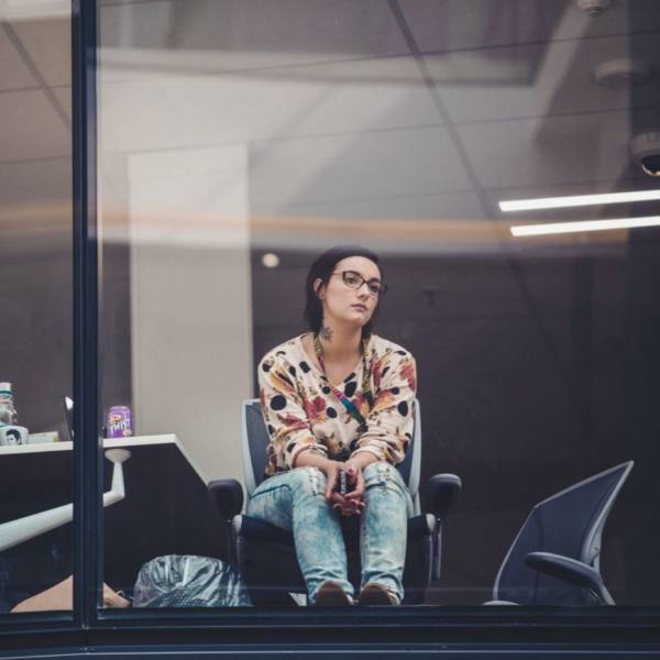 A young girl is sat in an office building. The camera is outside taking the picture through the window. She is thinking. She is wearing a blouse and jeans and appears to have been working. She has short brown hair and is wearing black rectangular glasses. She is looking off to the right side of the camera (from camera's view).