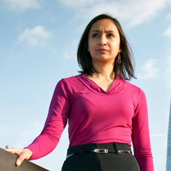 Roma Agrawal is looking out over the top of the camera. She is waring a pink top and a black skirt/trousers. She has shoulder-length, straight, brown hair and is high up as the background is a blue sky.