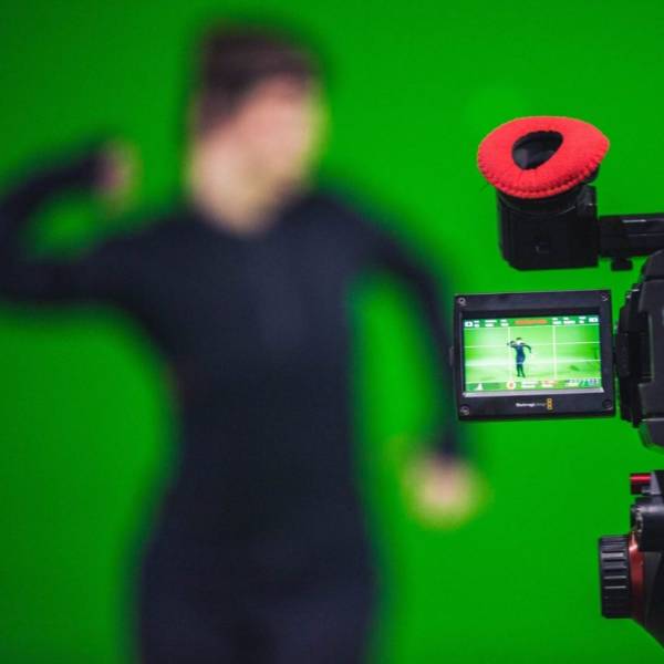 A lady is in front of a green screen wearing all black clothes. She appears to be dancing and can be seen clearly through a recording camera. The actual view of her is blurred.