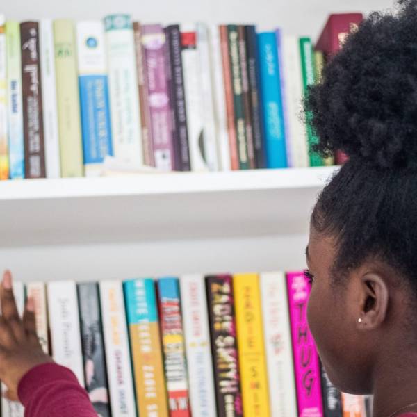 There is a young girl picking a book from a selection across two white shelves. The books are blurred as part of the background.