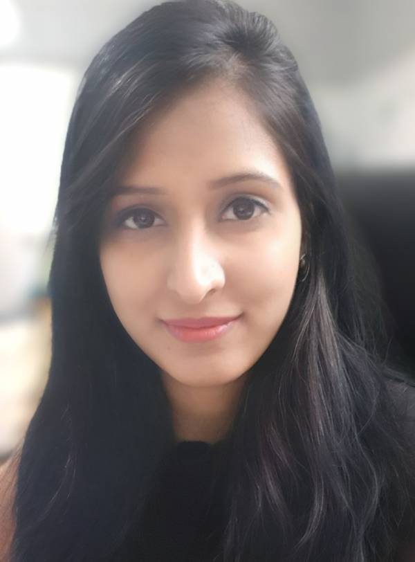 Anika Mahajan is smiling at the camera. She has long black hair and is wearing a brown t-shirt. She is sat at a desk however the background is blurred.