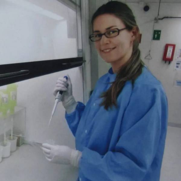 Ellen is in a lab. She is smiling at the camera. She is wearing a blue overall and a pair of white sterile gloves. She appears to be doing an experiment with a piece of equipment. She has long, brown hair tied back in a ponytail and is wearing black rectangular glasses.