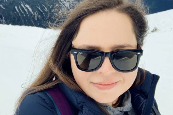 Karolina is smiling at the camera. She has long brown hair and is wearing a pair of sunglasses. She is also waring a blue coat and a grey jumper. Her hair has been blown around by the wind. She is stood on a snowy mountain.