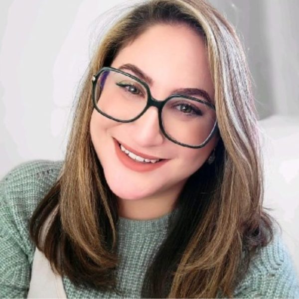 Julia is smiling at the camera. She has long blonde hair and is wearing black square glasses. She is sat in front of a white background. The picture repeats three times next to each other.
