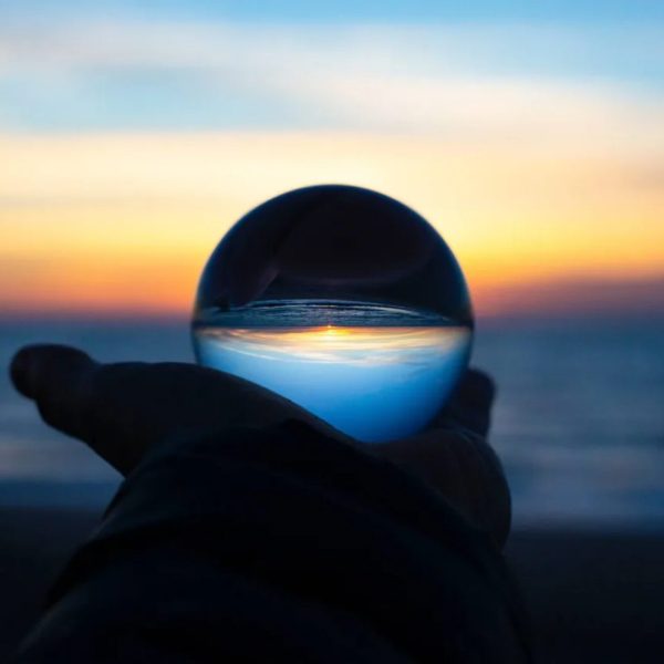 There is a hand outstretched holding a glass sphere. In the glass sphere, there is a reflected/inverted version of the beach at sunset, which is shown blurred in the background behind the sphere.