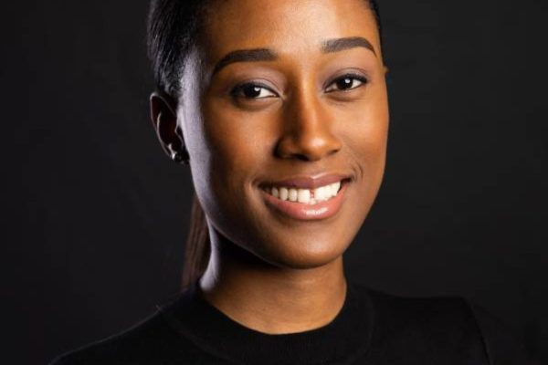 A young girl (Ifeoma Uzoegwu) is smiling at the camera. Her hair is in a pony tail and she is wearing a black top. She is stood in front of a black background. There are there of the same picture next to each other.