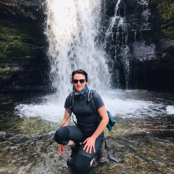 Sara from NewDay is smiling at the camera. She is wearing hiking gear and walking boots and is carrying a backpack. She is wearing sunglasses and a scarf around her neck. She is crouched down in front of a waterfall and it appears that she has walked there.