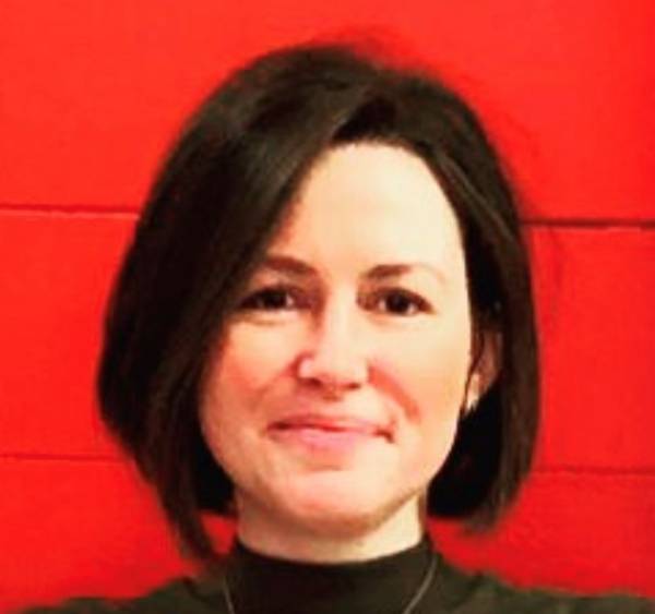 Sarah Grindall is smiling at the camera. She has short brown hair; it is styled in a bob with a parting down the right side (from camera's view point) She is wearing a black turtleneck top and is stood in front of a plain red background that appears to be made out of planks of wood.