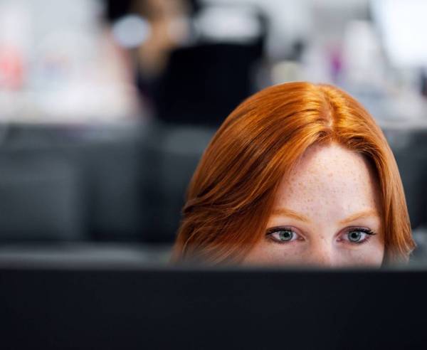 There is a girl working on a computer. Just hey eyes and the top of her head are visible over the top of the computer. She has ginger hair and green eyes. The background is blurred.