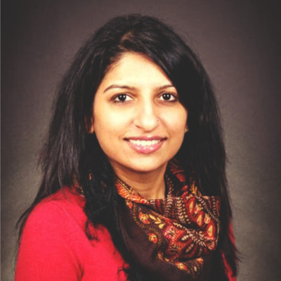 Lattika is looking at the camera smiling, she is wearing a black top with a red-pink cardigan. She has long black hair and is wearing a vibrantly coloured scarf around her neck. She is stood in front of a plain black wall.