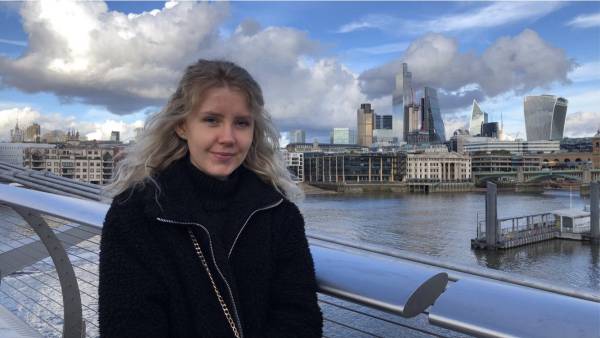 Charlotte from Nestle is smiling at the camera. She is standing on a bridge wearing a black coat with a silver zip. She has long, blonde, curly hair and is stood in front of a London horizon with skyscrapers visible on the right of the photo. The Thames is viible in the bottom half of the photo with a pier in the lower right hand side