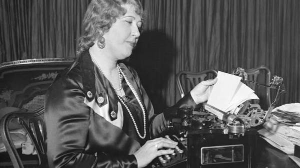 Beulah Henry is sat at a desk with a typewriter that she appears to be using, behind the typewriter is a stack of papers. She has short curly hair and is wearing several strings of pearls in varying lengths. The picture is in black and white and is taken in a old-fashioned room.